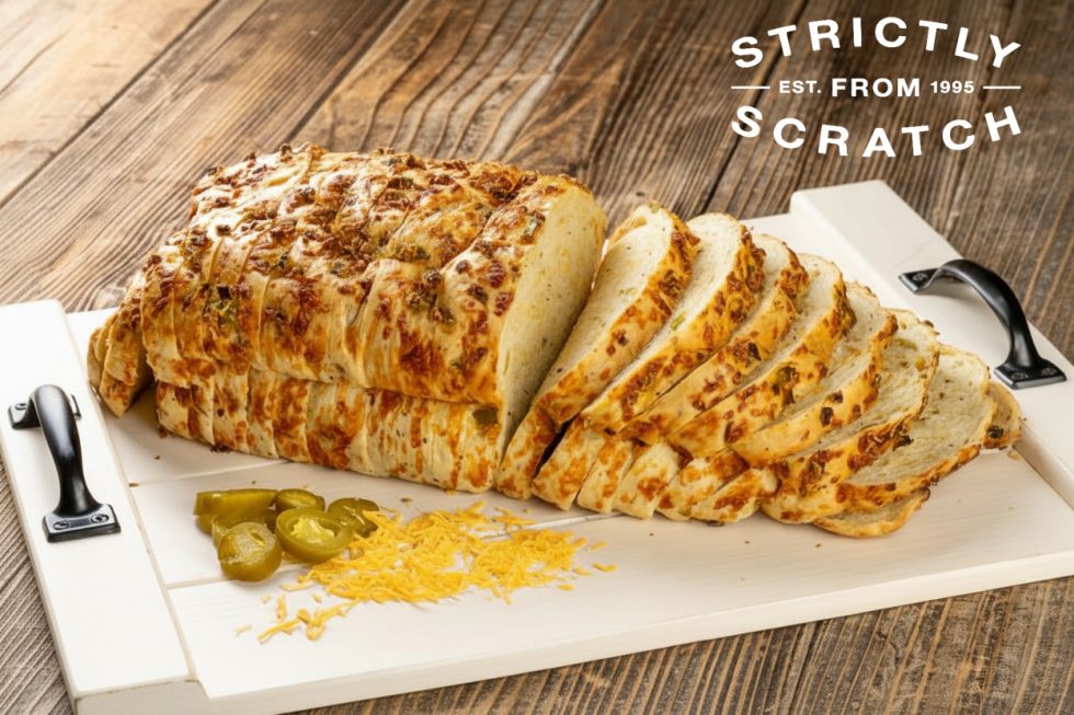 A sliced loaf of golden-brown Jalapeño Cheddar Pull-Apart Bread on a white cutting board, surrounded by fresh shredded cheddar cheese and pickled jalapeño slices on a wooden background.