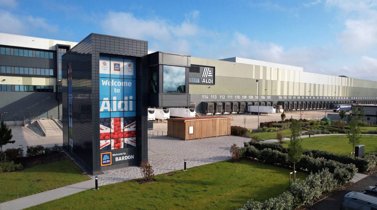Exterior of Aldi's Bardon Distribution Centre, a long gray warehouse with a "Welcome to Aldi" entrance tower, loading docks and landscaped grounds.