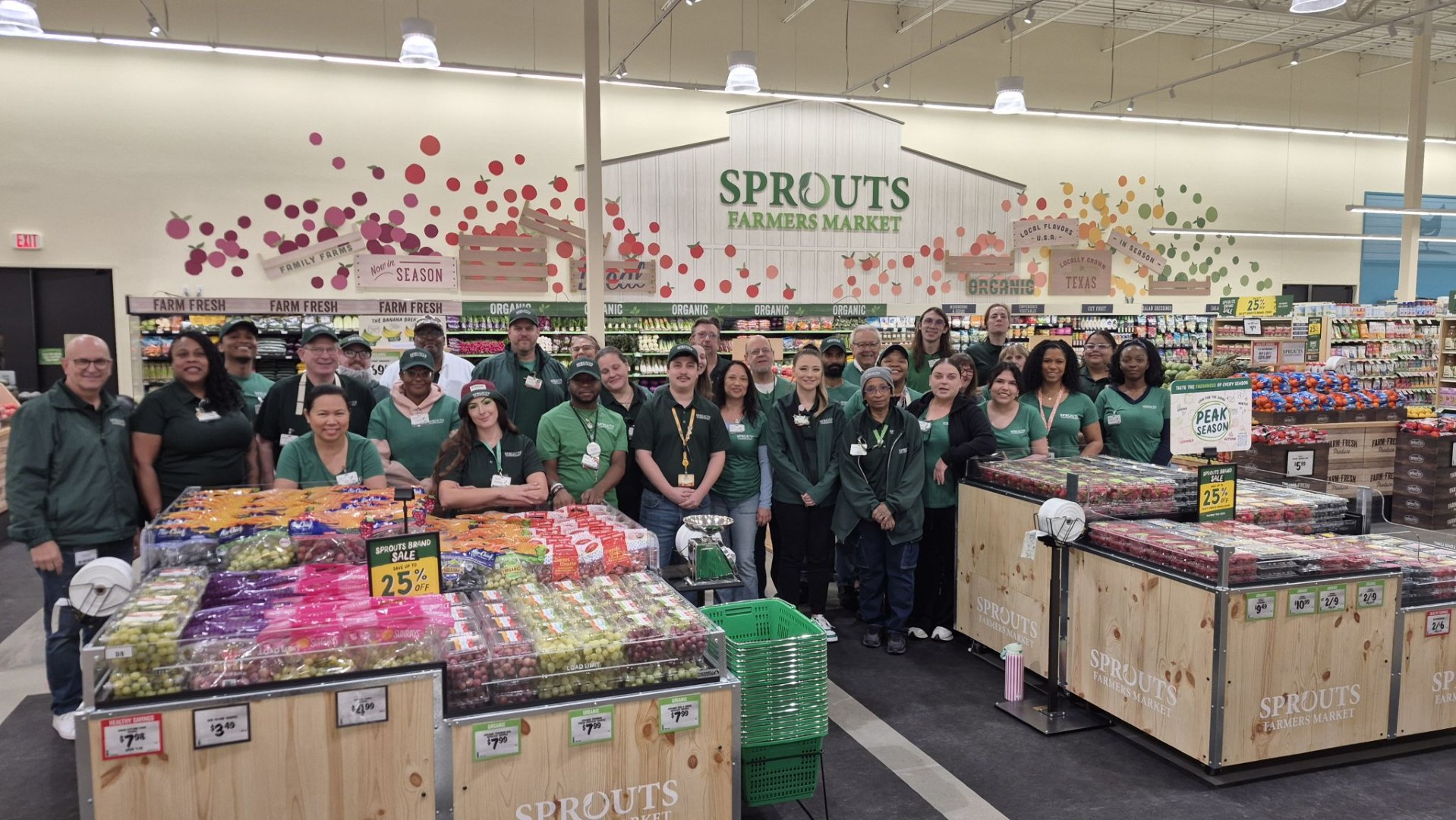 Sprouts Farmers Market employees in green shirts posing around wooden produce bins in a new store, with a large Sprouts Farmers Market sign on the wall behind them